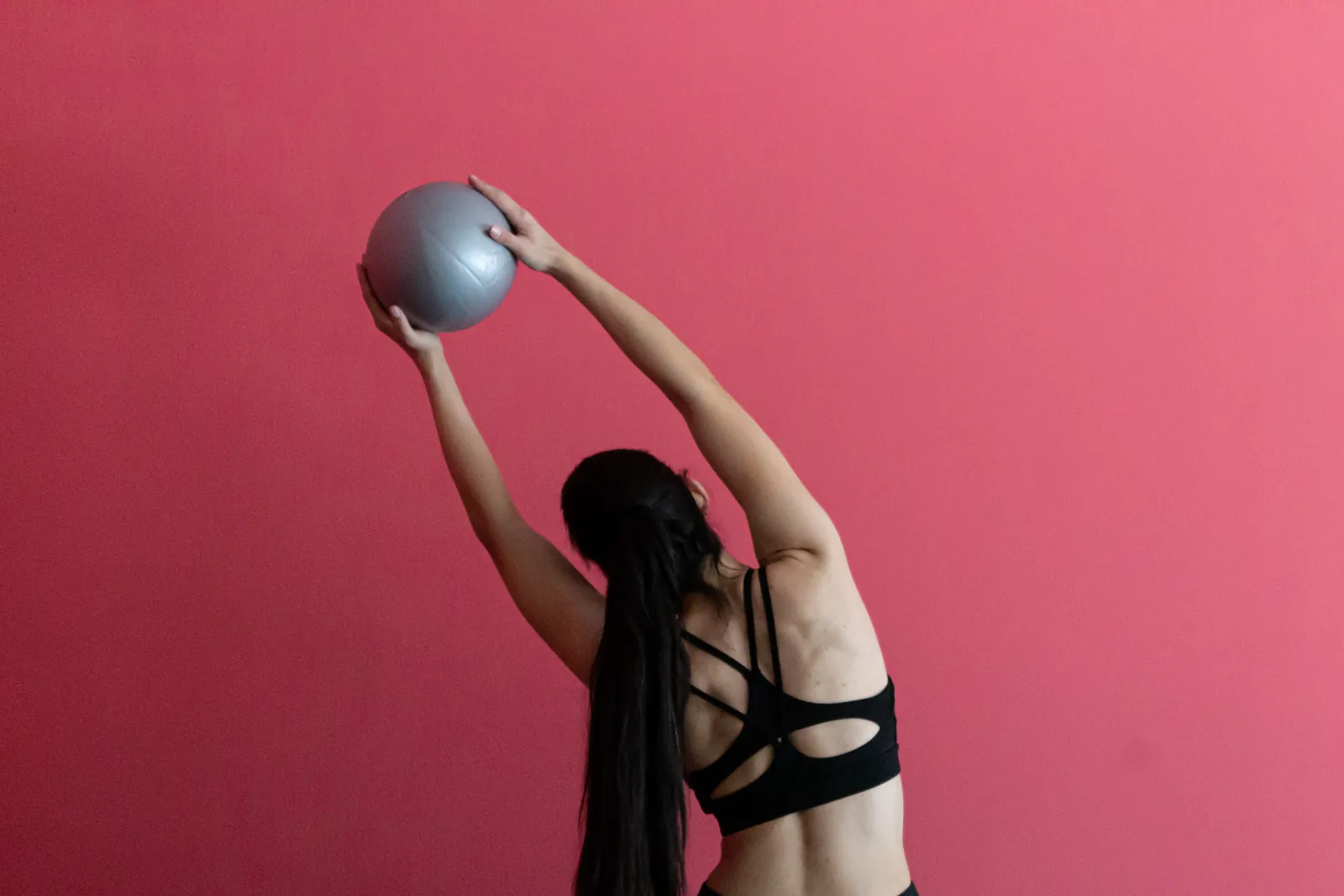 De espaldas, una mujer se estira hacia un lado con la pelota de Pilates frente al muro rosado del estudio Elevé.